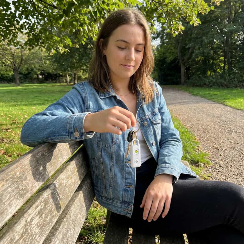 Personal safety alarm keychain in white, clipped to a set of keys, held on a park bench